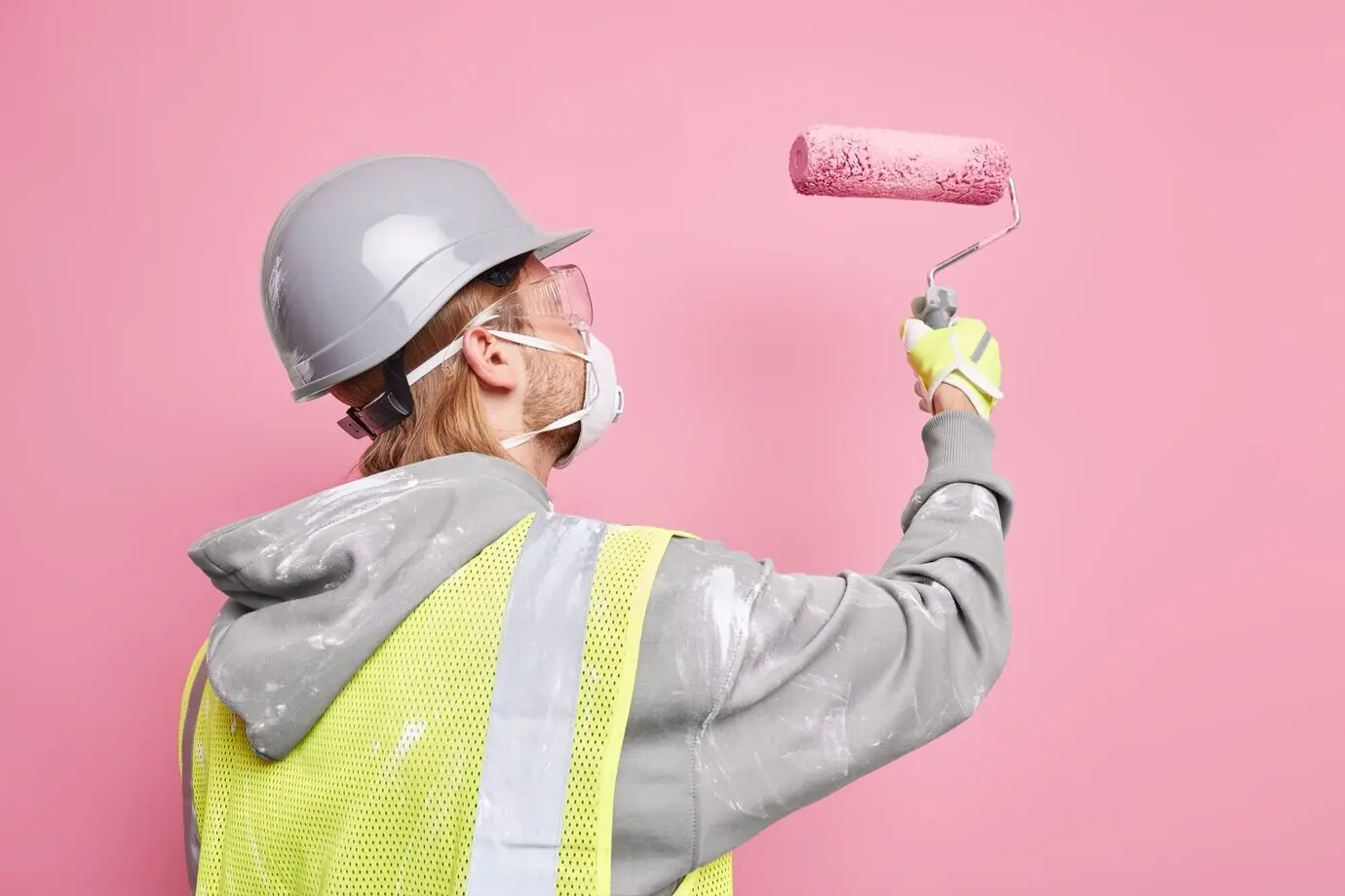 Side view of a busy handyman in a safety uniform, holding a paint roller and wearing a protective mask, working hard against a pink wall. Repair and renovation concept. Maintenance worker.