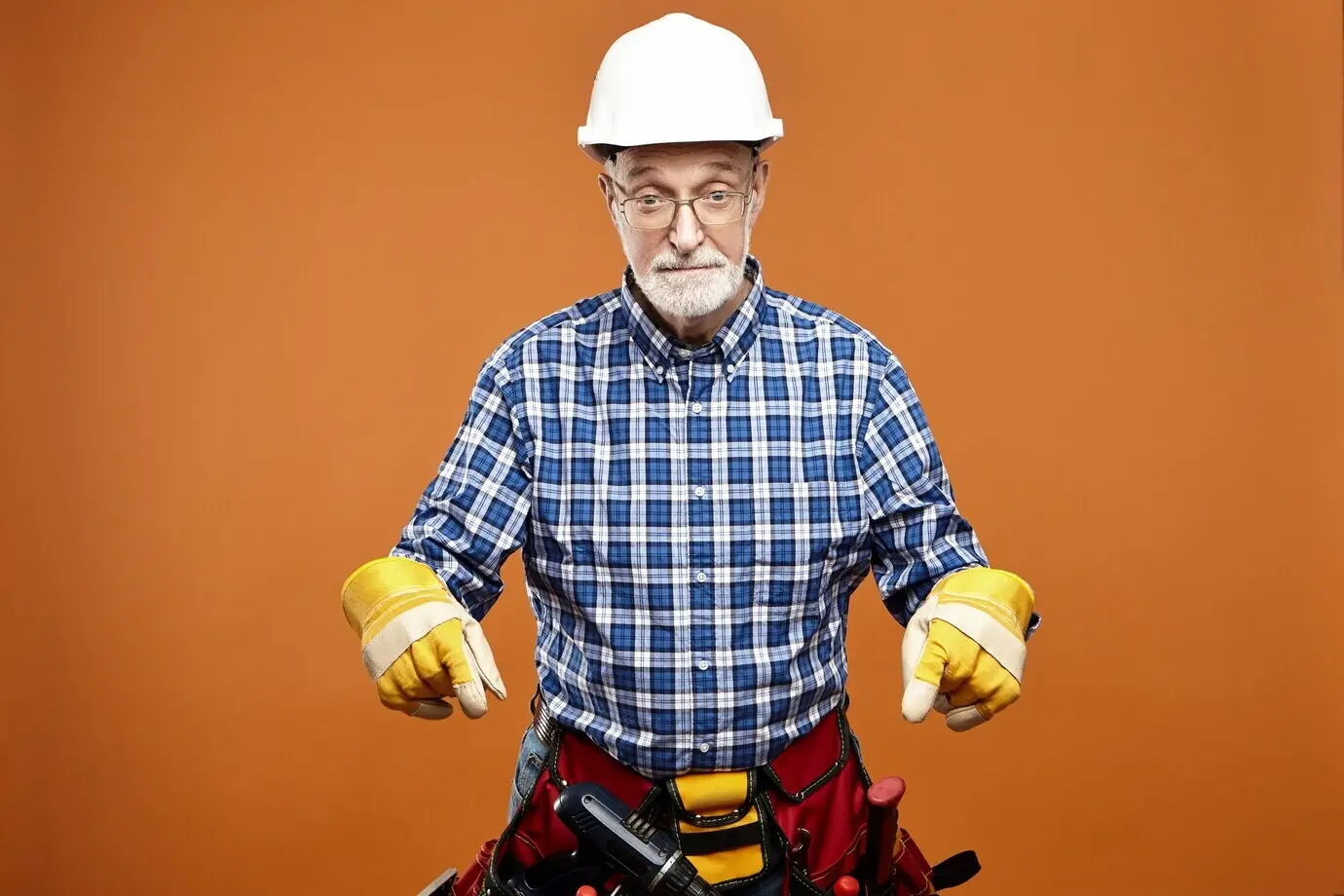 An emotional, funny elderly senior house builder wearing a protective helmet, safety gloves, and a bag of tools around his waist, with a shocked, surprised facial expression, pointing both index fingers downward.